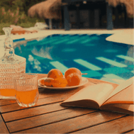 Naranjas, libro y bebida sobre una mesa frente a una piscina
